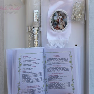 May include: A white baptism candle with a silver cross and chalice, a white satin ribbon with a religious image, and a small book with Spanish text about the Catholic faith.