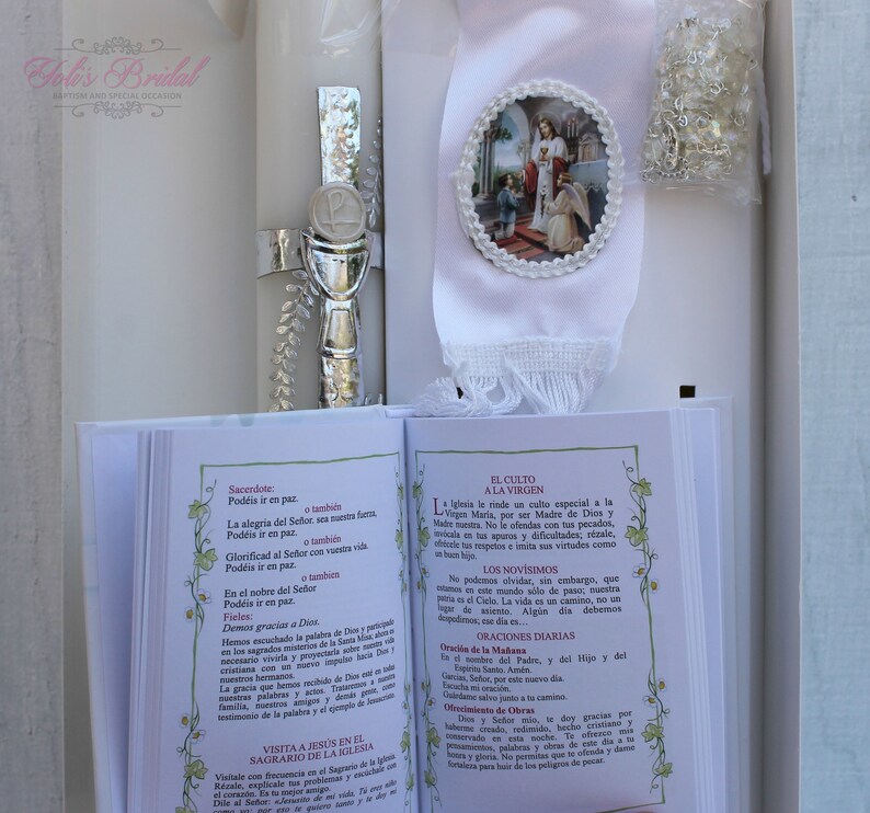 May include: A white baptism candle with a silver cross and chalice, a white satin ribbon with a religious image, and a small book with Spanish text about the Catholic faith.