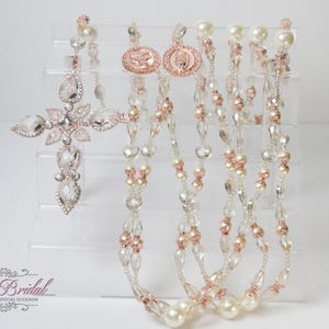 Può includere: Un set di tre collane di perle di cristallo e oro rosa con un grande ciondolo a croce di cristallo. Le collane sono esposte su un supporto in acrilico trasparente.