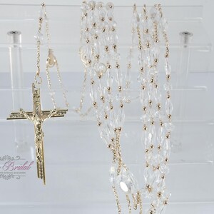 May include: A rosary featuring clear, faceted crystal beads interspersed with small gold-toned metal beads on a gold-toned chain. A gold-toned crucifix with a detailed figure of Jesus is attached to the end. The text Yoli's Bridal is visible.