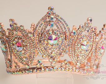 May include: A rose gold crown featuring iridescent rhinestones. The crown has a pointed top and a rounded base. It is adorned with a large, central rhinestone.