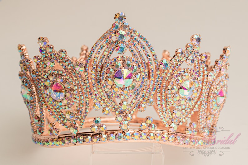 May include: A rose gold crown with iridescent rhinestones. The crown has a pointed top and a rounded base. It is decorated with a large, central rhinestone.