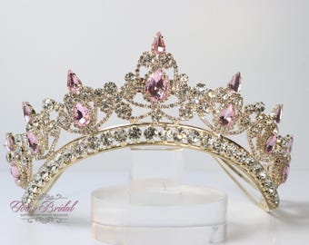 Puede incluir: Una tiara dorada con diamantes de imitación rosas y transparentes. La tiara tiene un diseño delicado con una corona puntiaguda y una base curva.