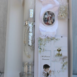 May include: A white gift box with a white satin ribbon and a silver-trimmed candle. The box also contains a white booklet with the text "Mi Primera Comuni&oacute;n" and a small white rosary.