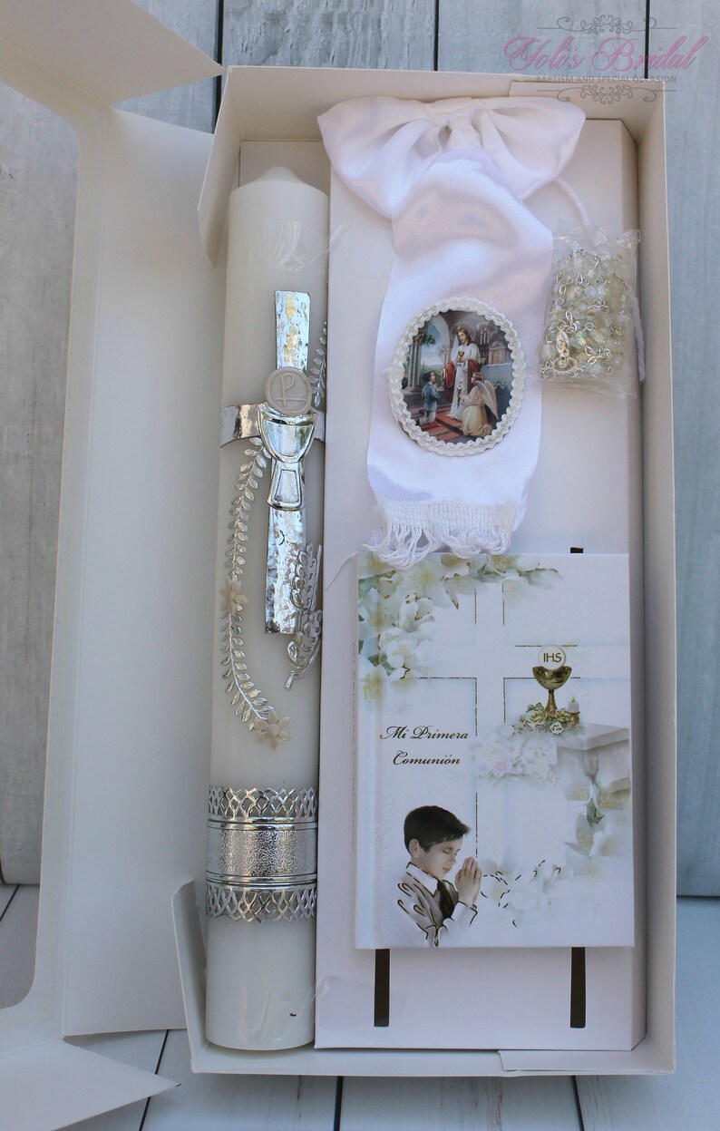May include: A white gift box with a white satin ribbon and a silver-trimmed candle. The box also contains a white booklet with the text "Mi Primera Comuni&oacute;n" and a small white rosary.