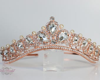 May include: A rose gold tiara with clear, sparkling crystals and faux pearls. The tiara features a curved band and is adorned with various sizes of crystals and small pearls. A clear stand supports the tiara.