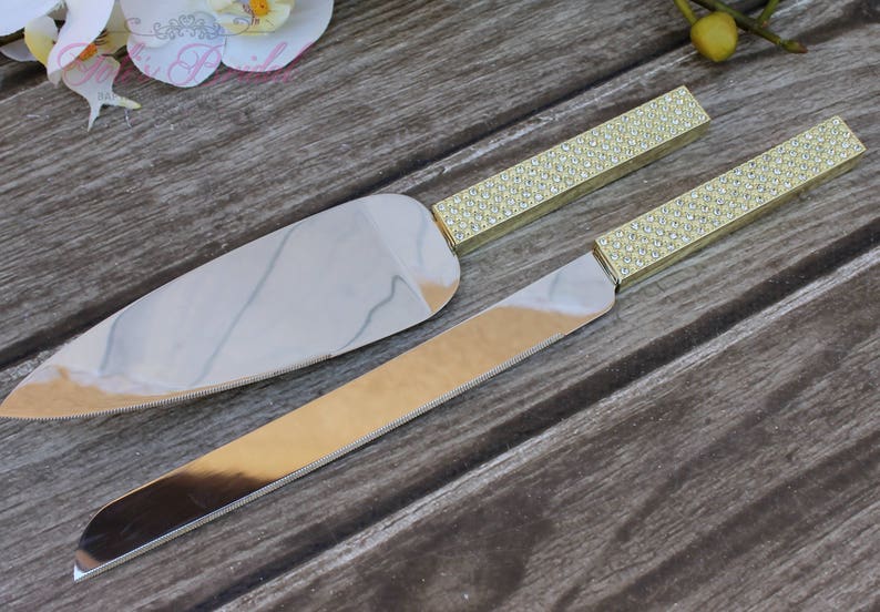 Puede incluir: Un juego de dos cuchillos para pastel de color dorado con mangos de diamantes de imitaci&oacute;n transparentes. Los cuchillos est&aacute;n sobre una superficie de madera.