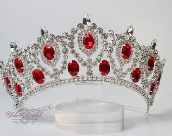 May include: Silver tiara with red gemstones and a delicate design. The tiara is on a white stand.