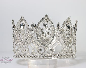 May include: A silver-toned crown with clear, sparkling rhinestones. The crown features a symmetrical design with multiple peaks and a base lined with smaller stones. The crown is likely for a special occasion, such as a wedding or pageant.