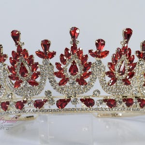 Op de afbeelding: Een goudkleurige kroon versierd met rode en heldere edelstenen. De kroon heeft een symmetrisch ontwerp met grote rode druppelvormige stenen en kleinere heldere stenen. Waarschijnlijk voor een speciale gelegenheid.