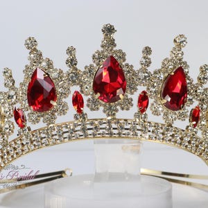 May include: A gold-toned crown with sparkling clear and red gemstones. The crown features large, teardrop-shaped red gems and smaller red and clear stones. The design is ornate and suitable for special occasions.