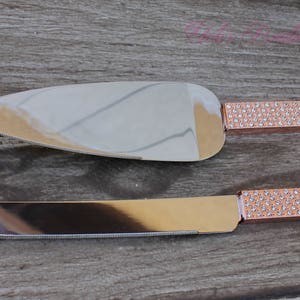 May include: A set of two cake knives with rose gold handles and clear rhinestone accents. The knives are on a wooden surface.
