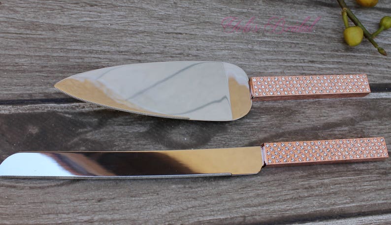 May include: A set of two cake knives with rose gold handles and clear rhinestone accents. The knives are on a wooden surface.