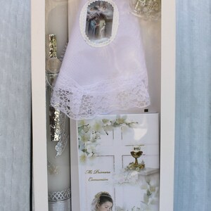 May include: A white box containing a white candle, a white lace doily, a white card with a picture of a girl praying, and a white rosary. The card has the text "Mi Primera Comuni&oacute;n" on it.