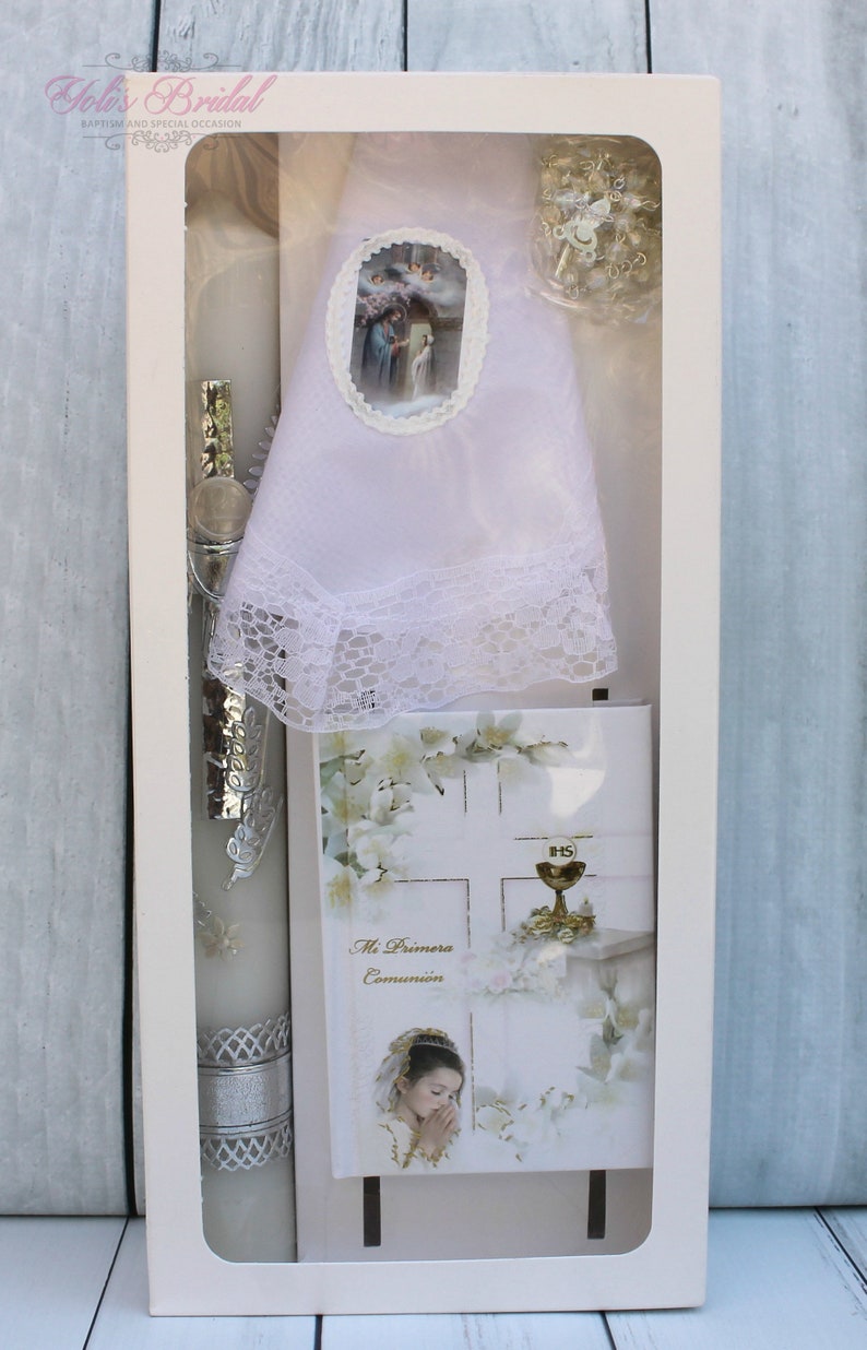 May include: A white box containing a white candle, a white lace doily, a white card with a picture of a girl praying, and a white rosary. The card has the text "Mi Primera Comuni&oacute;n" on it.
