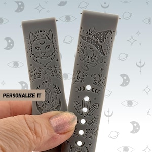 May include: Two gray watch bands with engraved designs. One features a cat with a crescent moon, the other a witch with a crescent moon hat. Both bands have floral, crystal, and celestial details. The text "PERSONALIZE IT" is visible.