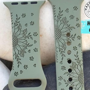 May include: Two sage green watch bands with engraved designs. Each band features a sunflower, leaves, and paw prints. The bands are made of a flexible material and have a classic watch band design.