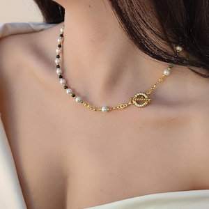 Op de afbeelding: Een goudkleurige ketting met witte parels en donkere kralen. De ketting heeft een gouden sluiting en wordt om de nek gedragen. Het sieraad is een ketting.