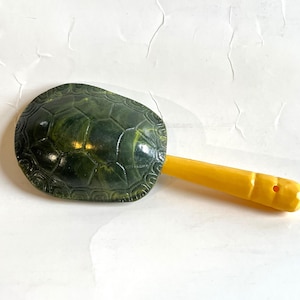 May include: A handheld rattle featuring a dark green, textured turtle shell top with a realistic scale pattern and a bright yellow, smooth, cylindrical handle with a small hole at the end, designed for producing sound when shaken.