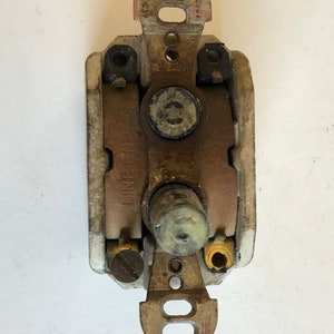 May include: A vintage electrical dimmer switch with a central knob and metal mounting brackets. The switch has a cream-colored body with visible wear and tear, and the metal components show signs of age and use.