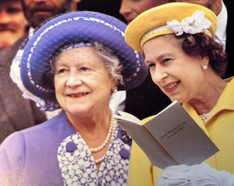May include: Two women smiling, one wearing a blue hat with a white trim and the other wearing a yellow hat with a white flower. One is holding a book with text.