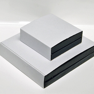 Może przedstawiać: Dwa białe pudełka z czarnymi obramowaniami ułożone jedno na drugim. G&oacute;rne pudełko jest nieco mniejsze od dolnego.