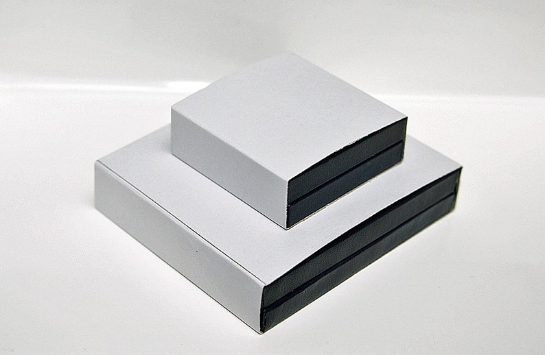 Może przedstawiać: Dwa białe pudełka z czarnymi obramowaniami ułożone jedno na drugim. G&oacute;rne pudełko jest nieco mniejsze od dolnego.