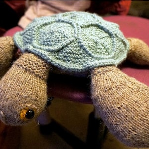 Op de afbeelding: Een gehaakt schildpad speelgoed met een lichtblauwe schil en bruine poten. De schildpad zit op een bruin oppervlak.