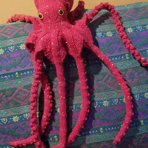 May include: A pink knitted octopus with yellow eyes. The octopus is sitting on a colorful patterned fabric.
