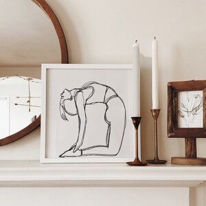 Könnte beinhalten: Ein weiß gerahmter Druck einer Linienzeichnung einer Person in einer Yoga-Pose. Der Druck hängt an einer Wand über einem weißen Kaminsims mit zwei Kerzenständern und einem Holzrahmen mit einem Druck darin.