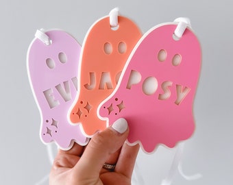 May include: Three colorful ghost-shaped tags in pink, orange, and lavender. Each tag has a name cut out: "EVI", "JAC", and "POSY". White ribbon is attached at the top. The tags are held against a light gray background.