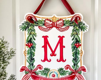 May include: A red and green Christmas door hanger with a gold frame and a large red letter "M" in the center. The hanger has a red ribbon and a white bow with a gold crown. The text "The Macalees" is on a red ribbon at the bottom.