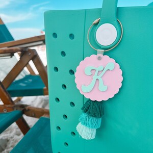May include: Teal tote bag with a pink and green tassel keychain. The keychain has a pink circle with a white letter K in the center.
