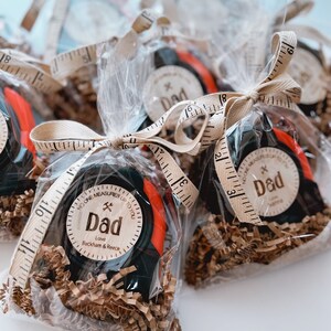 May include: A set of four small gift bags filled with brown shredded paper and a black and red tape measure with a wooden tag that reads "No one measures up to you Dad" with the text "Love Beckham & Reece".