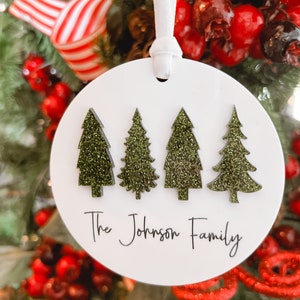 May include: White round Christmas ornament with four green glitter Christmas trees and the text "The Johnson Family".