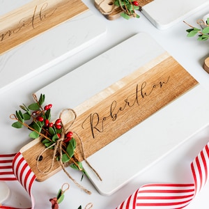 May include: Two white and wood cutting boards with the names "Smith" and "Robertson" engraved on them. The boards are decorated with sprigs of greenery and red berries, and a red and white striped ribbon.