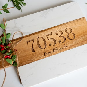 May include: A white marble and wood cutting board with the number 70538 and the text "franklin, la" engraved on the wood portion. The board is decorated with a sprig of greenery tied with twine.