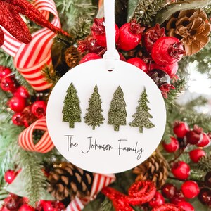 May include: White round Christmas ornament with four green glitter Christmas trees and the text "The Johnson Family".