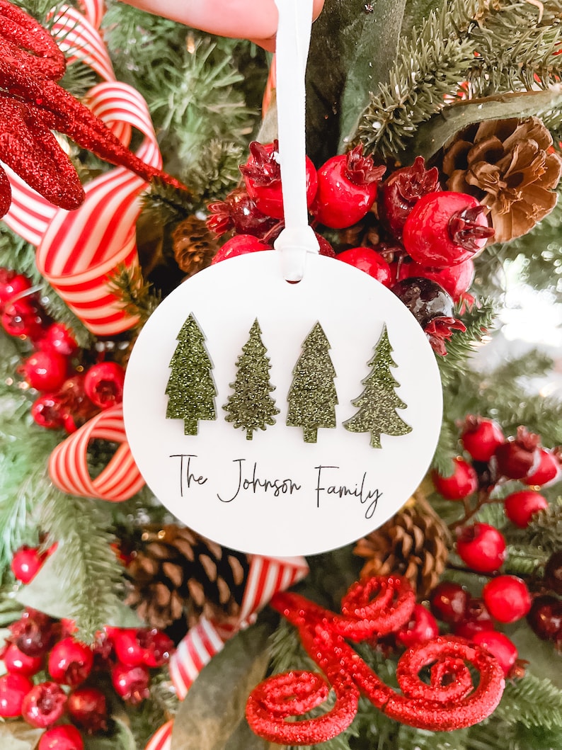 May include: White round Christmas ornament with four green glitter Christmas trees and the text "The Johnson Family".
