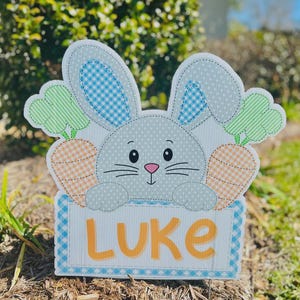 May include: A decorative Easter bunny yard sign with the name "LUKE" in orange letters. The bunny has blue gingham and white polka dot ears, with orange and green carrots on either side. The sign is made of wood.