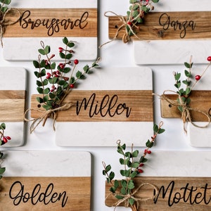 May include: Six white and brown marble serving boards with greenery and red berries. Each board has a different name written on the wood portion: Broussard, Garza, Peter, Miller, Golden, and Mottet.