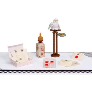 May include: A miniature Harry Potter-themed playset featuring a white owl perched on a brown stand, a bottle of ink, a box of letters, and wax seals. The set is arranged on a white surface with a black and white border.