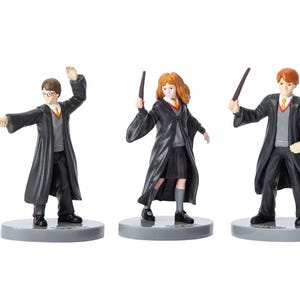 May include: Three figurines of characters from a fantasy series. Each figure wears a black robe, gray sweater, and a yellow and red tie. They are holding wands and standing on gray circular bases.