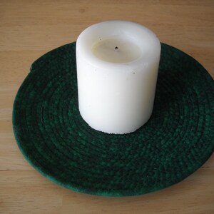 May include: A white pillar candle sits on a dark green, circular woven coaster. The candle is unlit and has a slightly uneven top. The coaster is made of a tightly woven material, creating a textured surface.