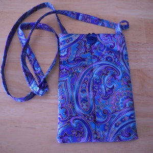 May include: A small, rectangular crossbody bag with a vibrant paisley pattern in shades of blue and purple. The bag has a long strap and a button closure. The fabric has a detailed, swirling design.