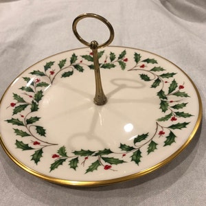 May include: A two-tiered serving tray with a white background and a gold rim. The tray is decorated with a holly and berry wreath design. The handle is gold-coloured.