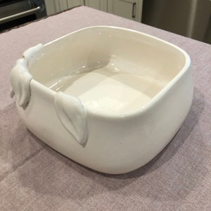 Könnte beinhalten: Eine quadratische, cremefarbene Keramikschale mit abgerundeten Ecken. Die Schale hat eine glatte, glänzende Oberfläche und ist mit blattartigen Verzierungen auf einer Seite versehen. Die Schale ist leer und steht auf einer hellvioletten Oberfläche.