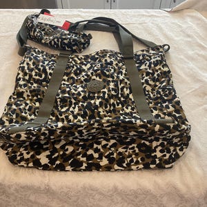 May include: A large, brown and white leopard print tote bag with green straps and a Kipling logo tag. The bag has a zipper closure and two top handles.
