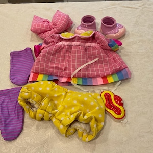 May include: A collection of baby doll clothes, including a pink plaid dress with a rainbow trim, a yellow polka dot diaper cover, two pairs of purple striped socks, a pink bow, and pink shoes.