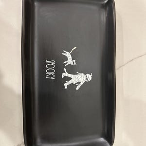May include: Black rectangular ceramic tray with rounded corners. The tray features a white graphic of a mummy and a cat, with the word "SPOOKY" in white vertical text. The tray is likely for Halloween.